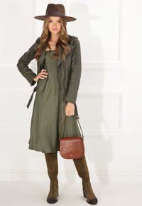 Olivgrünes Kleid, braune Lederjacke, brauner Filzhut, knielange Wildlederstiefel und eine braune Leder-Schultertasche. Glatte Texturen und geschichtetes Design.