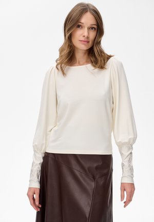 Langärmliges cremefarbenes Oberteil mit rundem Ausschnitt und puffärmel; verfügt über bestickte Akzente an den Bündchen; kombiniert mit einem braunen Lederrock.