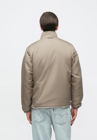 Brun polstret jakke i letvægtsstof med høj krave, ribbet manchetter og kant, glat tekstur, båret over lyseblå denimjeans.