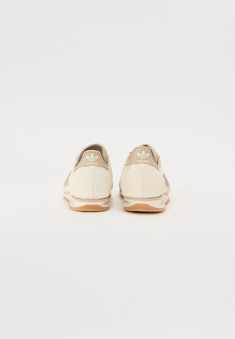 Baskets de sport beiges avec une texture lisse, des panneaux latéraux en maille et une semelle en caoutchouc. Marque visible sur le talon et la languette.