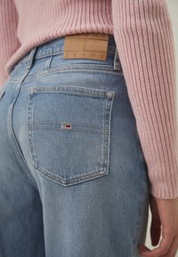 Svijetloplave traperice s tamnosmešnim kožnim patčem sa natpisom "TOMMY JEANS" na stražnjoj strani. Imaju dva stražnja džepa s šivenim detaljima.