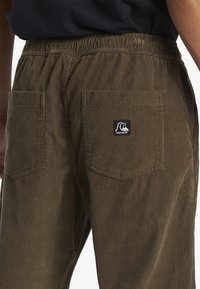 Brown corduroy pants with a ribbed texture, featuring an elastic waistband and two back pockets. Includes a logo patch on one pocket.