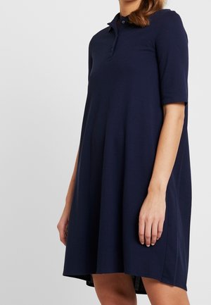 Žena oblečená v volně střižených námořnicky modrých polokabátových šatech s krátkými rukávy na jednobarevném světlém pozadí.