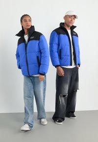 To modeller bærer blå og sorte pufferjakker. Jakkerne har en lynlås foran, kontrasterende farver og er kombineret med løse jeans og sneakers.