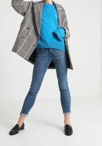 Schwarz-weiß karierten Doppelreiher-Mantel über einem leuchtend blauen Pullover, kombiniert mit blauen Skinny-Jeans und schwarzen Loafers.