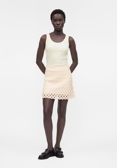 Γυναίκα όρθια με τα χέρια πίσω από την πλάτη, φορώντας ένα αμάνικο, ανοιχτό κίτρινο τοπ, μια κρεμ δαντελωτή μίνι φούστα και μαύρα παπούτσια Mary Jane.