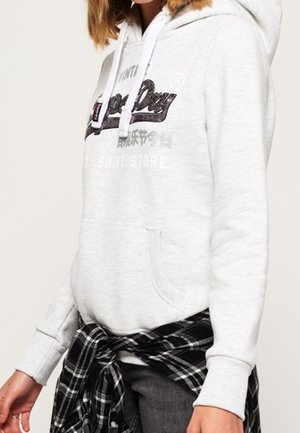 Personne portant un sweat à capuche gris clair avec un logo noir et blanc, des cordons blancs, et une chemise à carreaux noir et blanc nouée autour de la taille.