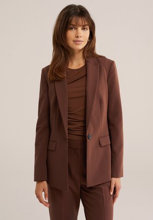Femme aux cheveux bruns et raides, portant un blazer marron, un pantalon assorti et un haut marron, debout devant un fond clair uni.