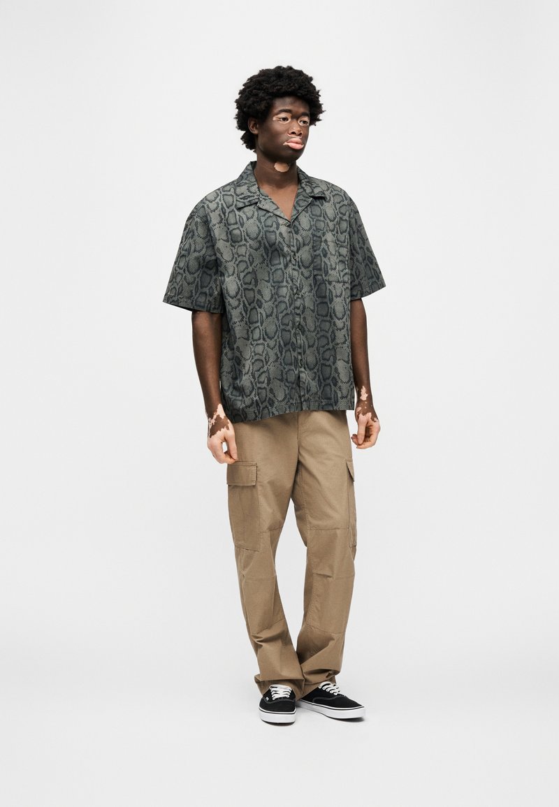 Camicia a maniche corte con stampa di serpente verde scuro, vestibilità rilassata, abbinata a pantaloni cargo beige con tasche laterali e sneakers nere.