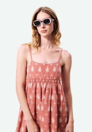 Femme aux cheveux blonds mi-longs portant des lunettes de soleil noires à monture blanche et une robe rose sans manches avec des motifs floraux blancs.