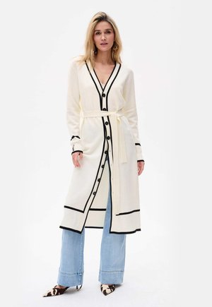 Langer cremfarbener Cardigan mit schwarzem Saum, Knöpfen und Gürtel-Detail. Getragen über hellblauen weiten Jeans und gemusterten Absätzen.