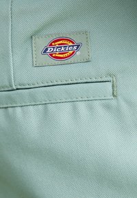 Lichtgroene stof met een zichtbaar Dickies merkpatch. De patch heeft een cirkelvormig logo met rode, gele en blauwe kleuren.
