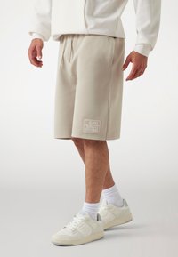 Beige cotton blend shorts with an elastic waistband, side pockets, and a patch featuring "GUESS" and "1981" on the left leg. White sneakers.