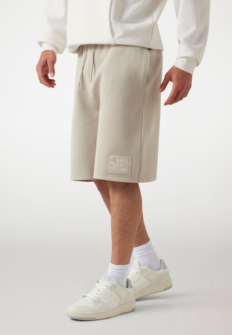 Beige cotton blend shorts with an elastic waistband, side pockets, and a patch featuring "GUESS" and "1981" on the left leg. White sneakers.