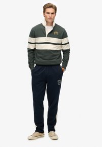 Bosgroene pullover met witte strepen en logo, gecombineerd met marineblauwe joggers met een logo en beige accenten. Casual schoeisel inbegrepen.