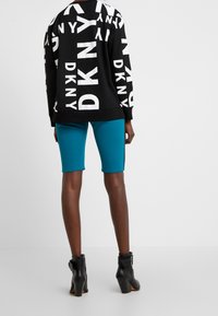Persoon met zwarte enkellaarsjes, teal-kleurige korte broek tot aan de knie en een zwart sweatshirt met grote witte "DKNY" tekst vanaf de achterkant.
