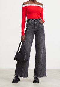 Nainen, jolla on yllään olkaimeton punainen toppi, jossa on valkoiset Tommy Jeans -reunat, korkeavyötäröiset, leveät mustat farkut, mustat saappaat ja musta käsilaukku.