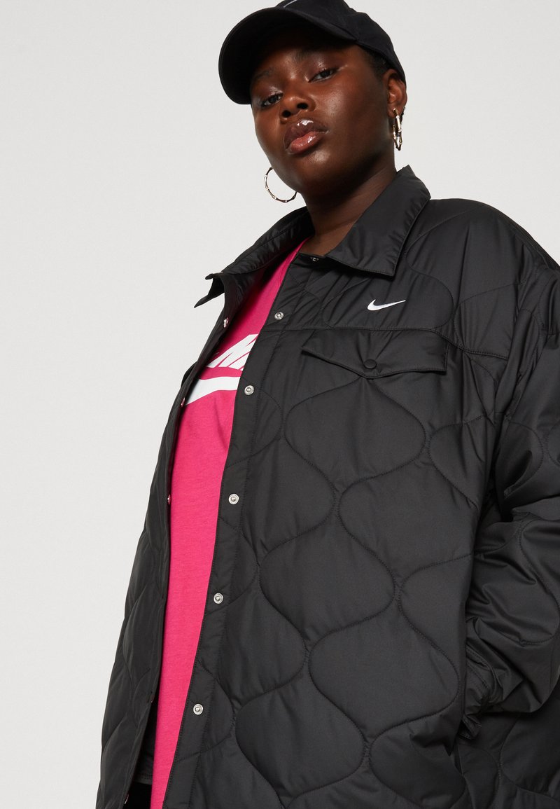 Veste noire matelassée avec une fermeture à boutons-pression, dotée d'une poche sur la poitrine et d'un petit logo blanc. Portée par-dessus un t-shirt graphique rose.