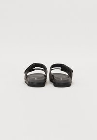 Black sandals with a flat sole, featuring a padded toe post and adjustable Velcro strap. Smooth texture with a minimalist design.