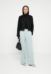 Jachetă scurtă neagră cu textură peste un guler înalt negru, asortată cu pantaloni evazați de culoare bleu pal și poșetă neagră, evidențiată de pantofi cu vârf ascuțit.