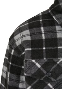 Chemise en flanelle polaire à motif à carreaux noir et gris avec des accents blancs. Comprend une poche à bouton et un col classique. Texture douce.