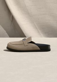 Μπεζ σουέντ μοκασίνι τύπου slip-on με ασημένια αγκράφα και μαύρη σόλα, εκτεθειμένο σε ανοιχτή επιφάνεια με φόντο υφή από μπεζ ύφασμα.