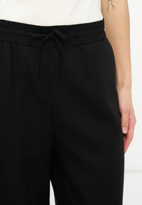 Κοντινή λήψη μαύρων παντελονιών με ελαστική μέση και κορδόνι, φορεμένα με λευκό τοπ; εμφανές χέρι με ελάχιστο τατουάζ με γραμμή στο πάνω μέρος του αντιβραχίου.