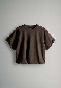 Brūna īsām piedurknēm T-kreklu ar apaļu kakla izgriezumu, brīvu piegriezumu un mīkstu kokvilnas audumu. Tam ir nedaudz pūkainas piedurknes un īsāks dizains.