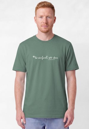 Homme aux cheveux roux portant un t-shirt vert avec un texte en allemand "So einfach ist das", debout devant un fond blanc uni.