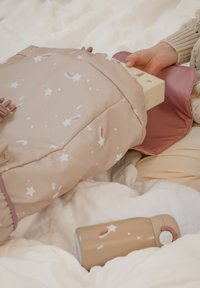 Zachte, beige rugzak met sterpatroon en roze accenten, gedeeltelijk open om een lichtgekleurde doos binnen te laten zien; een bijpassende fles ligt op wit stof.