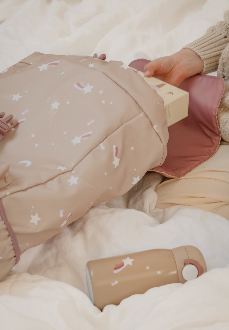 Zachte, beige rugzak met sterpatroon en roze accenten, gedeeltelijk open om een lichtgekleurde doos binnen te laten zien; een bijpassende fles ligt op wit stof.