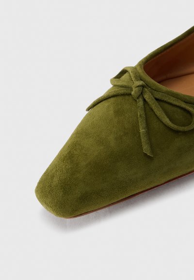 Επίπεδο παπούτσι από σουέτ σε ελιά πράσινο χρώμα με μυτερή μύτη και διακοσμητικό φιόγκο στην κορυφή, εμφανίζοντας μέρος της ταμπά εσωτερικής επένδυσης.