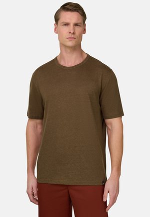 Mann mit kurzen Haaren, der ein schlichtes braunes kurzärmeliges T-Shirt und rostfarbene Hosen trägt, steht vor hellem Hintergrund.