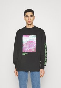 Čierne tričko s long-sleeve, na prednej strane s farebnou grafickou potlačou a zelenými textovými akcentmi na rukávoch. Tkanina vyzerá mäkko.