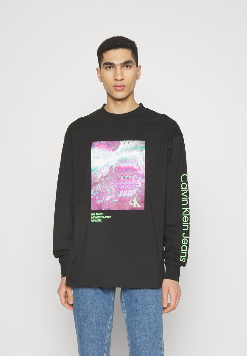 Čierne tričko s long-sleeve, na prednej strane s farebnou grafickou potlačou a zelenými textovými akcentmi na rukávoch. Tkanina vyzerá mäkko.