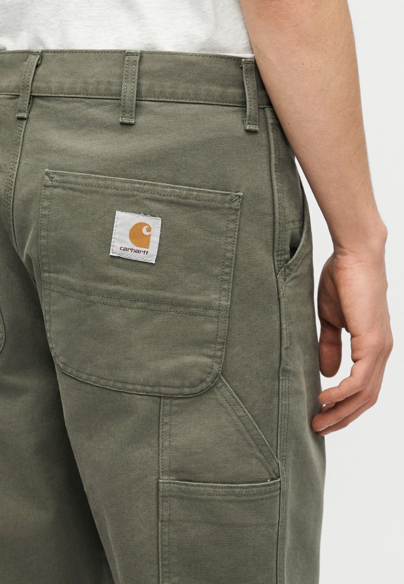 Person trägt olivgrüne Carhartt-Hose mit großer Gesäßtasche und Logo-Patch, Hand entspannt seitlich neben dem Körper vor weißem Hintergrund.