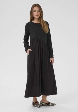 Langes schwarzes Kleid mit eng anliegendem Oberteil, ausgestelltem Rock, langen Ärmeln und Taschen. Kombiniert mit schwarz-weißen gestreiften Ballerinas.