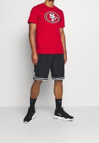 Rode katoenen t-shirt met "SF" logo, gecombineerd met zwarte shorts met witte zijstrepen en zwarte sport sneakers.