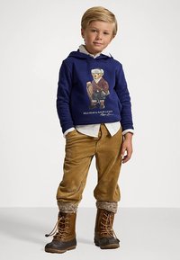 Jūras spēku kapuci ar lāča grafiku, gaiši brūnas korduroja bikses un brūni šņorējami zābaki ar vilnas akcentiem. Zem tā balta krekla apkakle.