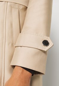 Gros plan sur la manche beige d'un manteau avec un bouton noir à quatre trous sur une sangle réglable, montrant une partie de la main et la couture de la poche.