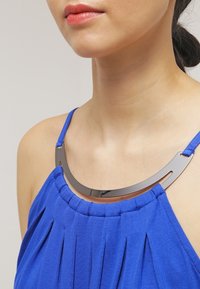 Haut plissé bleu avec des bretelles fines et un collier métallique courbé et brillant reposant sur le décolleté. Texture de tissu lisse, design minimaliste.