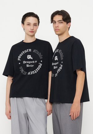 Dvaja modeli nosiaci zhodné čierne oversized tričká s kruhovým bielym textom a sivé široké nohavice, stojaci na jednoduchom bielom pozadí.