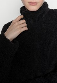 Schwarzer Fleecepullover mit hohem Kragen und weicher, strukturierter Oberfläche. Verfügt über einen Knopfverschluss am Kragen und breite, einlagige Bündchen.