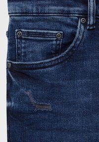 Gros plan sur la poche d'un jean en denim bleu foncé avec des rivets visibles et une petite déchirure horizontale usée sous la poche.