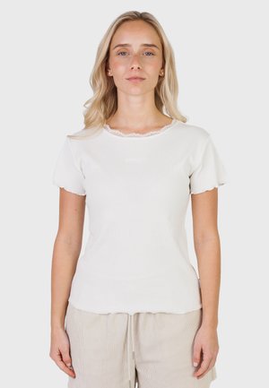 Blondynka w białej bluzce z krótkim rękawem ozdobionej koronką i beżowych spodniach, stojąca na jednolitym, jasnym tle.