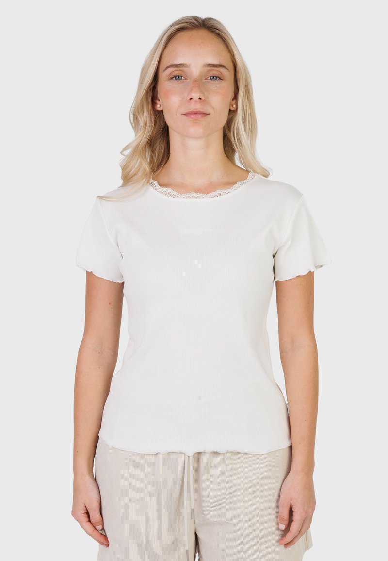 Femme blonde portant un haut blanc à manches courtes avec une bordure en dentelle et un pantalon beige, debout devant un fond clair uni.