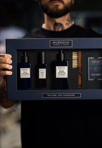 Gåvoset med tre mörkblå flaskor märkta för skäggvård: fuktighetskräm, olja och schampo, tillsammans med en träborste i en elegant låda.