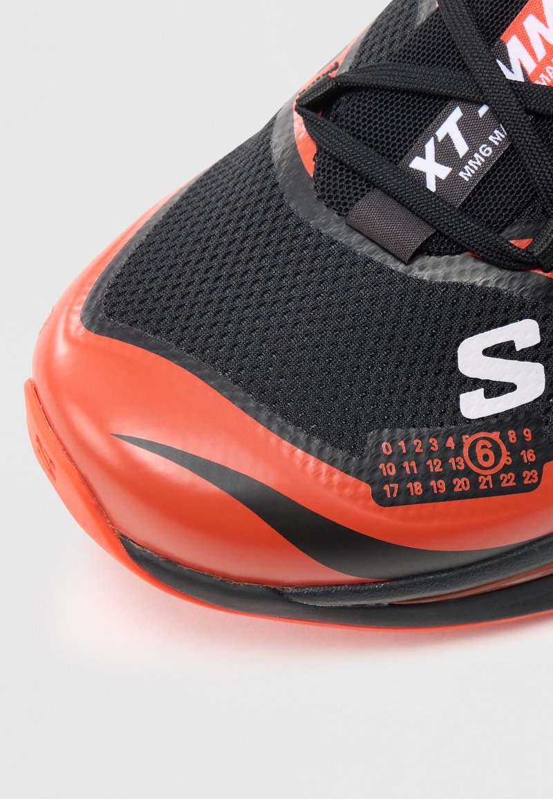 Sort og orange sneaker med en tekstureret mesh-overdel, glat gummitå og en nummereret skala på siden til størrelsesangivelse.