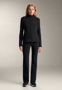 Czarna sportowa kurtka z wysokim kołnierzem i pełnym zamkiem, w zestawieniu z dopasowanymi spodniami. Gładki materiał i elegancki design. Czarne sneakersy dopełniają całość.