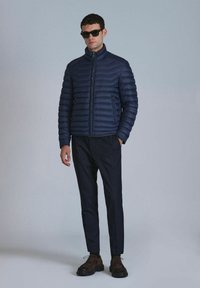 Navy puffer jacket with quilted design, high collar, and front zipper, paired with dark trousers and brown lace-up shoes.